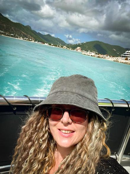 Live cam girl Aubrey Sky takes a selfie on a boat on a turquoise sea with a hilly green landscape and harbour in the background