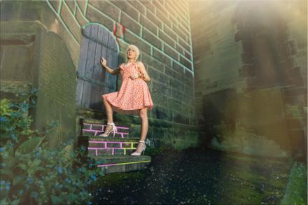 Goddess Alimony wears a red and white polka dot summer dress and pink heels as she climbs outdoor stone steps in a church graveyard heading toward a wooden castle door