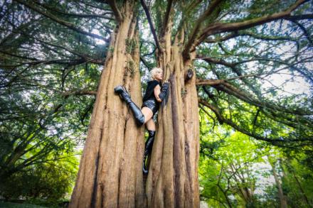 Goddess Alimony poses stuck between two trees staring off into the distance wearing black PVC outfit and boots