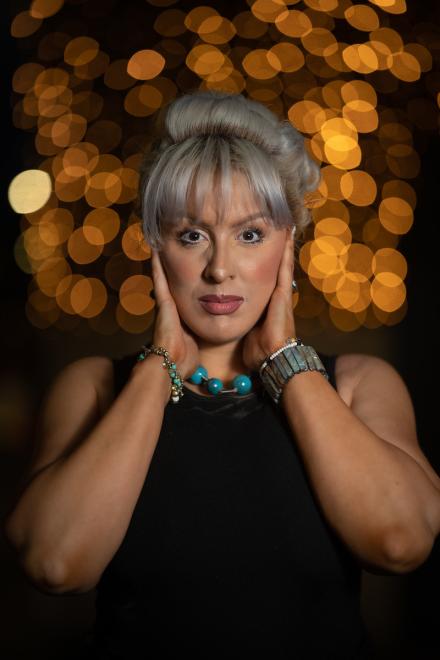 Goddess Alimony stands in front of distant fairy lights at night with her blonde hair up wearing a black dress with both hands on the sides of her face