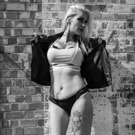 Jesi Storm standing up against a wall outdoors in a white top leather jacket and black panties holding the jacket open