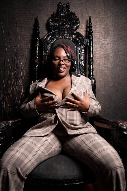 Oshun Waters sits wearing a suit and glasses on a black throne while pushing her big tits up
