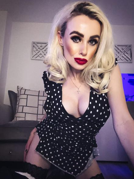 Blonde live cams model Syren Sexton kneels in front of a grey sofa wearing black dress with white polka dots and black stockings