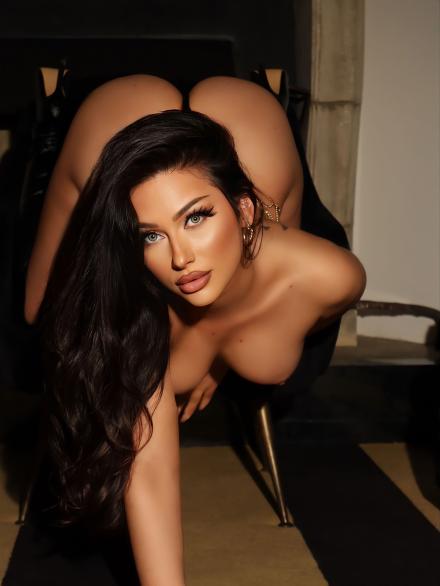 Aria Rose looks into the camera leaning forward off a chair with arm on the ground and big ass showing in the background