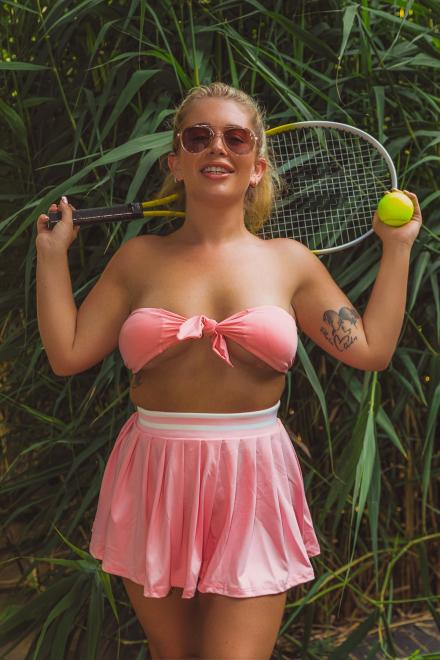 Darcy May stands front and smiling in front of a big green busy wearing pink bikini top, miniskirt and sunglasses while holding a tennis racket and ball behind her neck Darcy May stands front and smiling in front of a big green busy wearing pink bikini top, miniskirt and sunglasses while holding a tennis racket and ball behind her neck